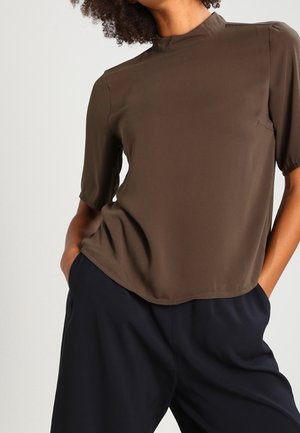 Woman wearing a brown high-neck short-sleeve top and loose black pants with hands in pockets against a plain background.