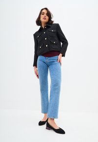 Giacca nera corta con bottoni dorati e quattro tasche frontali, abbinata a jeans svasati blu chiaro e tacchi neri con accenti bordeaux.