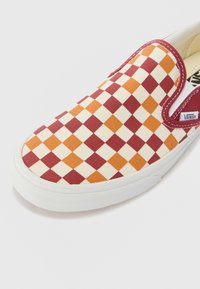 Slip-on sneaker with red, orange, and cream checkered pattern. Features a white rubber sole, elastic side accents, and Vans branding.