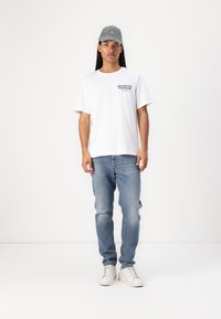 White cotton t-shirt with "REPUBLIQUE FRANCAISE" printed in black, paired with light blue jeans and gray cap, worn with white sneakers.