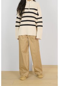 Personne portant un pantalon beige à jambes larges, des mocassins couleur noisette, et un pull en tricot crème avec des rayures horizontales noires et un col à demi-zip.