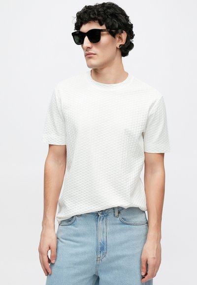 Λευκό T-shirt με υφή και σχέδιο ρόμβου, στρογγυλή λαιμόκοψη, κοντά μανίκια, συνδυασμένο με ανοιχτόχρωμο τζιν σε μπλε και μαύρα γυαλιά ηλίου.