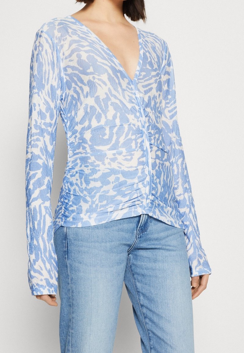 Hellblaue Bluse mit langen Ärmeln und V-Ausschnitt, mit einem weißen abstrakten Muster und gerafften Seitendetails für Form und Textur.