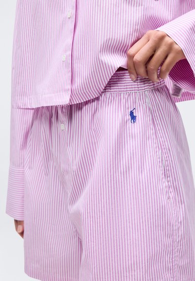 Χέρι που προσαρμόζει ροζ και λευκά ριγέ σορτς με ελαστική μέση και μπλε κεντημένο λογότυπο, συνδυασμένα με πουκάμισο με κουμπιά σε ίδιο σχέδιο.