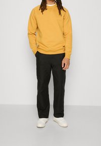 Persona in piedi che indossa un felpa giallo senape, pantaloni neri e sneakers bianche su uno sfondo grigio chiaro uniforme.