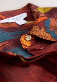 Red and orange patterned shirt with a collar, featuring a gold button and textured fabric. Label visible beneath the collar.