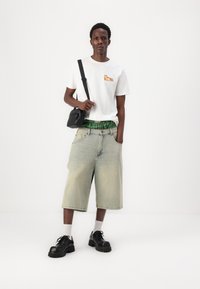 Λευκό μπλουζάκι με πολύχρωμο γραφικό, τζιν σορτς, μαύρα χοντρά παπούτσια και μικρή μ�αύρη τσάντα. Ορατός πράσινος καρό ιμάντας.