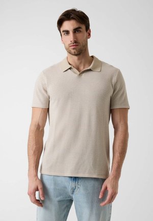 Mann mit kurzen braunen Haaren, der ein beiges, gestreiftes Poloshirt und hellblaue Jeans trägt, steht vor einem schlichten, hellen Hintergrund.