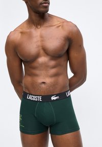 Άνδρας χωρίς πουκάμισο, με σταυρωμένα χέρια, φοράει σκούρα πράσινα μποξεράκια Lacoste με μαύρη μέση και λευκό λογότυπο σε απλό φόντο.