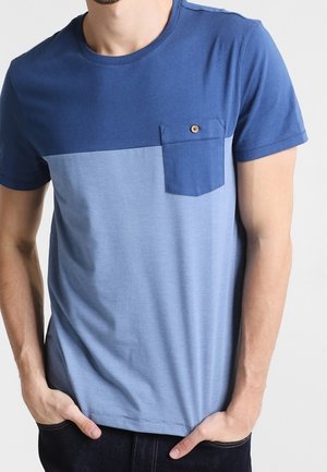 Mężczyzna w niebieskiej koszulce typu color-block z krótkim rękawem i guzikową kieszenią na piersi, z rękami w kieszeniach ciemnych spodni.