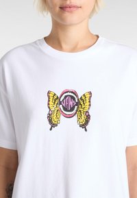 Hvid t-shirt med et centralt graphic af gule og sorte sommerfuglevinger, der omkranser en pink mund med "VANS" i fed sorte bogstaver.