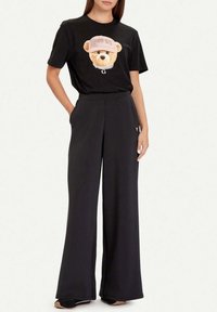 Camiseta negra con un gráfico de un oso con una gorra rosa y texto "GUESS", combinada con pantalones negros de pierna ancha y un pequeño detalle de logo.