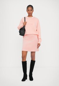 Sweatshirt rosa com logo em relevo, combinado com uma saia em veludo cotelê rosa e botas altas de camurça pretas. Acessório em forma de bolsa preta.
