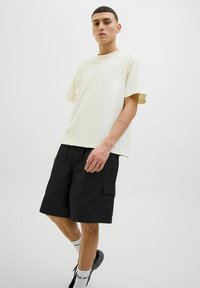 Tricou din bumbac de culoare galben deschis, cu mâneci scurte, purtat împreună cu pantaloni scurți cargo negri, cu buzunare. Modelul poartă sosete albe și pantofi negri.