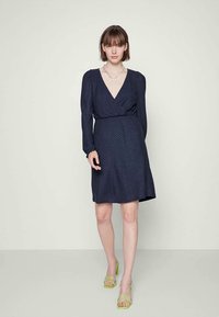 Mulher vestindo um vestido azul-marinho até ao joelho, com mangas longas e decote em V, combinado com sandálias de tiras verde-lima e um colar de corrente.