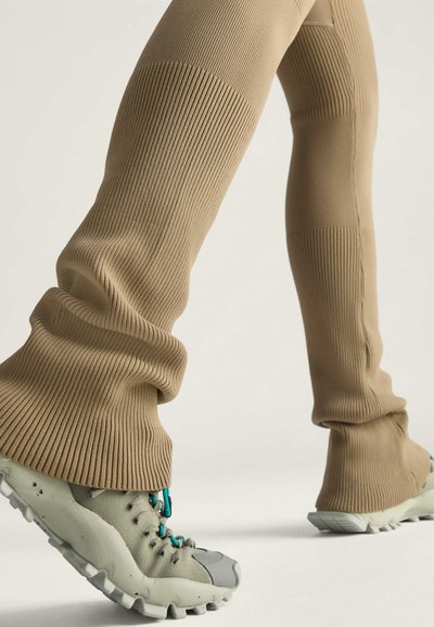 Bež hrpane tajice s opuštenim krojem, uparenih s sivim i tirkiznim sportskim cipelama koje imaju teksturiranu potplate i detaljan sustav vezivanja.