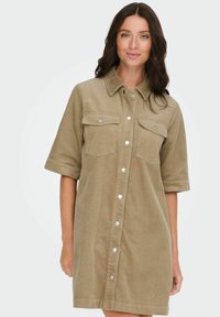 Haljina košulja od kežualnog flanela boje kaki s kopčanjem na gumbe sprijeda, dva džepa na prsima, kratkim rukavima i opuštenim krojem.