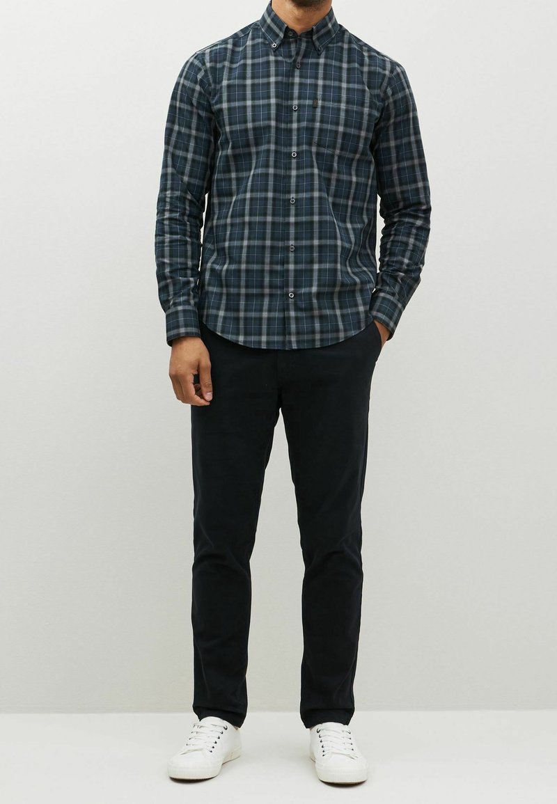 Homme portant une chemise à carreaux bleu foncé et vert, un pantalon noir et des baskets blanches, debout avec une main dans la poche.