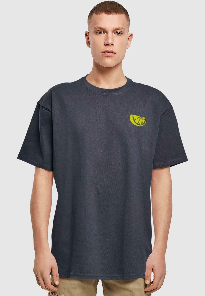 Mørkegrå bomuld t-shirt med løs pasform, der har et lille grønt limegrafik på venstre bryst. Korte ærmer og rund halsudskæring.