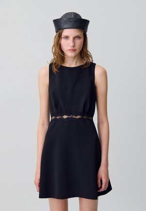 Femme portant une robe noire sans manches avec un détail en dentelle à la taille et un chapeau de marin en cuir noir, se tenant devant un fond gris uni.