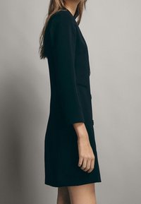 Schwarzes maßgeschneidertes Kleid mit langen Ärmeln, strukturiertem Schnitt, Ein-Knopf-Verschluss und Seitentaschen, das über eine glatte Stofftextur verfügt.