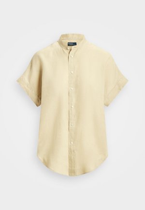 Beiges kurzärmeliges Hemd mit Stehkragen, abgerundetem Saum und sichtbaren Nähten vor hellem, einfarbigem Hintergrund.