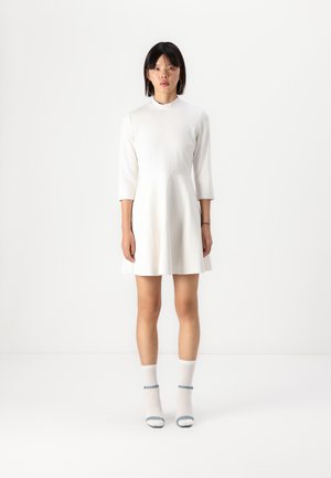 Robe blanche à manches courtes avec un col haut, jupe évasée, texture lisse, associée à des chaussettes blanches et des chaussures à talons avec accents bleus.