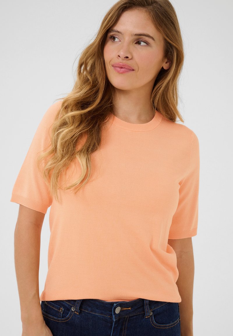 Pull en tricot à manches courtes et col rond de couleur orange clair. Texture douce avec ourlet et poignets côtelés. Associé à un jean en denim bleu foncé.