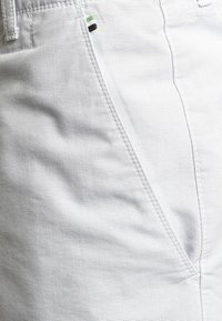 Shorts gris clair en tissu texturé, avec une poche latérale ornée d'un petit accent de couleur vert et des détails de couture noirs.