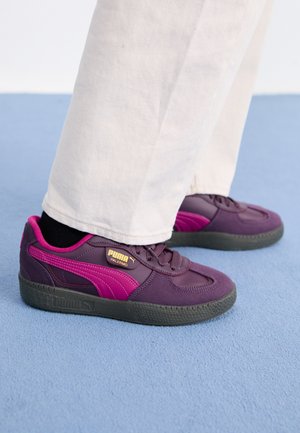 Purple and pink sneaker with rubber sole, featuring a textured surface, laces, and gold branding on the side. White pants contrast the shoes.
