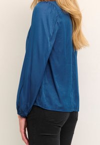 Femme portant un chemisier bleu brillant à manches longues, associé à un jean noir ajusté, vue de profil sur un fond uni.