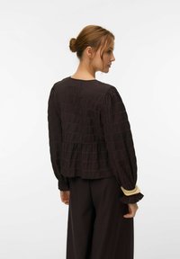 Kvinne med hår satt opp i en knute, iført en teksturert mørk bluse med lange ermer og matchende løse bukser, vist fra ryggen og siden mot en hvit bakgrunn.