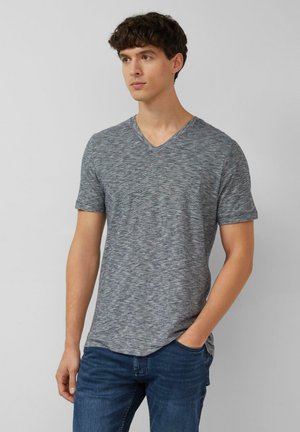 Jonge man met een grijs V-hals T-shirt met korte mouwen en een blauwe spijkerbroek, staand met één hand in de zak, kijkt iets naar de zijkant.