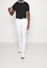 Svart polo skjorta med krage som har vita ränder, i kombination med vita byxor och vita sneakers, håller en golfklubba.