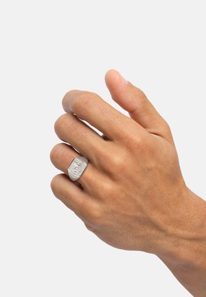 Silberner Ring mit rechteckiger Form und eingraviertem Design, getragen an einer Hand mit mittlerem Hautton. Glatte Metalloberfläche, minimalistischer Stil.