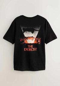 Melnā kokvilnas t-kreklā ir grafiskais dizains ar sarkanām un baltām krāsām, kas ietver tekstu "DEMONU IZBĀDĪTĀJS" un attēlus, kas saistīti ar šo tēmu.