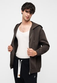 Sweat à capuche marron à fermeture éclair avec une texture douce, doté d'une grande capuche et de poignets côtelés, porté sur un débardeur blanc et un pantalon de survêtement noir avec des cordons blancs.