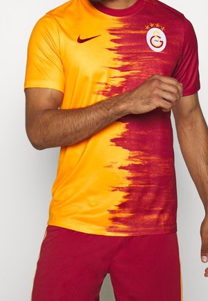 Man draagt een voetbalshirt van Galatasaray in bordeauxrood en geel met het clublogo en de Nike-swoosh, gecombineerd met bordeauxrode shorts.