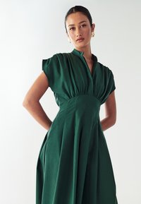 Žalia kelių ilgiai suknelė, pagaminta iš minkštos, tekstūruotos medžiagos, su mandarinų apykakle, trumpomis pūstomis rankovėmis ir vertikaliomis pūtimis sijone.