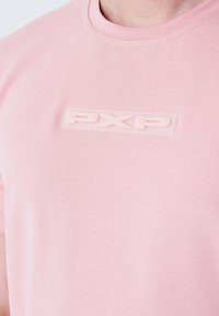 Camiseta de manga corta de color rosa claro, hecha de algodón suave, con un logo "PXP" en relieve de goma en el centro. Textura suave, escote clásico.