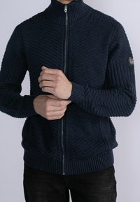 Mörkblå zippered tröja med ribbad hög hals, texturerade diagonala mönster och åtsittande ärmmuddar. Har sidofickor och logotyppatch.