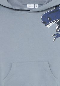 Felpa per bambini di colore azzurro chiaro con tasca frontale e una grafica parziale di un dinosauro che mostra denti e artiglio sul lato destro dell'upper chest.