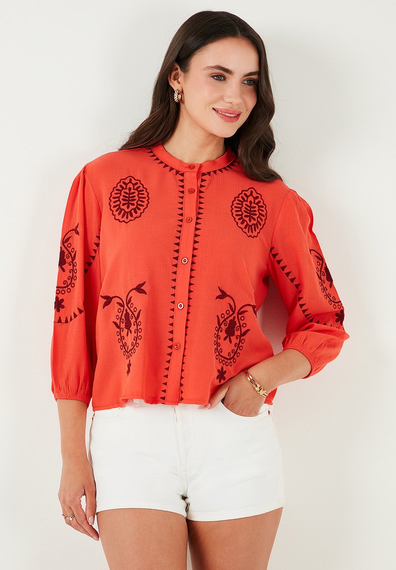 Orangenbluse mit schwarzer Stickerei, die Blumen- und geometrische Muster zeigt. Sie hat einen runden Kragen, eine Knopfverschlüsse und Dreiviertelärmel.