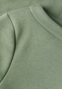 Zelená bavlněná tkanina zblízka, která ukazuje hladkou strukturu, žebrovaný výstřih a uvolněný, zaoblený tvar s jemnými detaily šití.