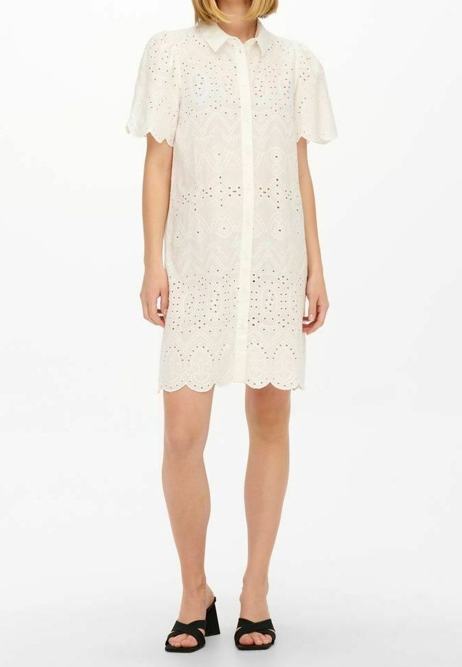 Femme portant une robe blanche en dentelle œillet à manches courtes avec un ourlet festonné et des sandales à talons ouvertes noires, debout sur fond blanc.