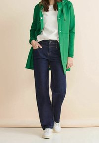 Giacca lunga verde, top bianco con trama, jeans blu scuro a gamba larga e sneakers bianche. La giacca presenta un cappuccio e chiusure a scatto.
