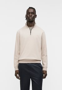 Μπεζ φούτερ με φερμουάρ, ψηλό γιακά, πλεκτά μανίκια και μικρό λογότυπο στο στήθος, συνδυασμένο με σκούρο, άνετο παντελόνι.