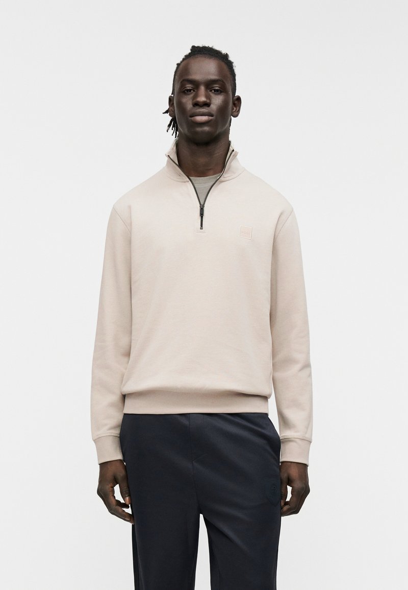 Μπεζ φούτερ με φερμουάρ, ψηλό γιακά, πλεκτά μανίκια και μικρό λογότυπο στο στήθος, συνδυασμένο με σκούρο, άνετο παντελόνι.