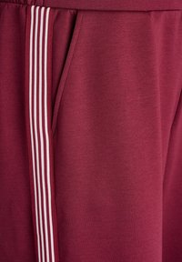 Burgunderfarbene Jogginghose aus weichem Stoff, mit weißen dreifachen Streifen an der Seite, einem elastischen Bund und einer Seitentasche.