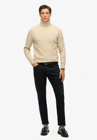 Jersey de punto beige con cuello alto y un acabado texturizado, combinado con unos vaqueros azul oscuro y mocasines negros. Diseño simple, sin patrones visibles.